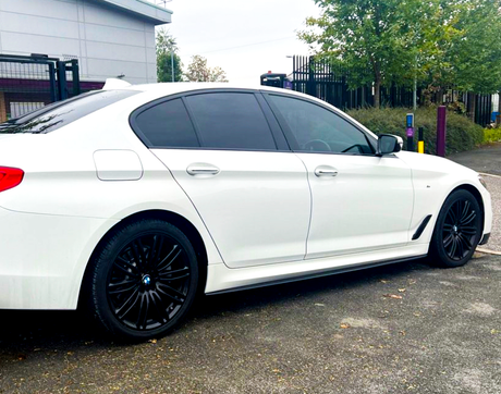A white sedan with tinted windows and black wheels is parked on a paved surface. It is near a fenced area with trees and an industrial building in the background.