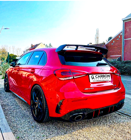 A red Mercedes-Benz parked on gravel, displaying a rear spoiler and dual exhausts, beside brick houses. License plate reads: "CARBON ACCENTS, WWW.CARBONACCENTS.CO.UK."