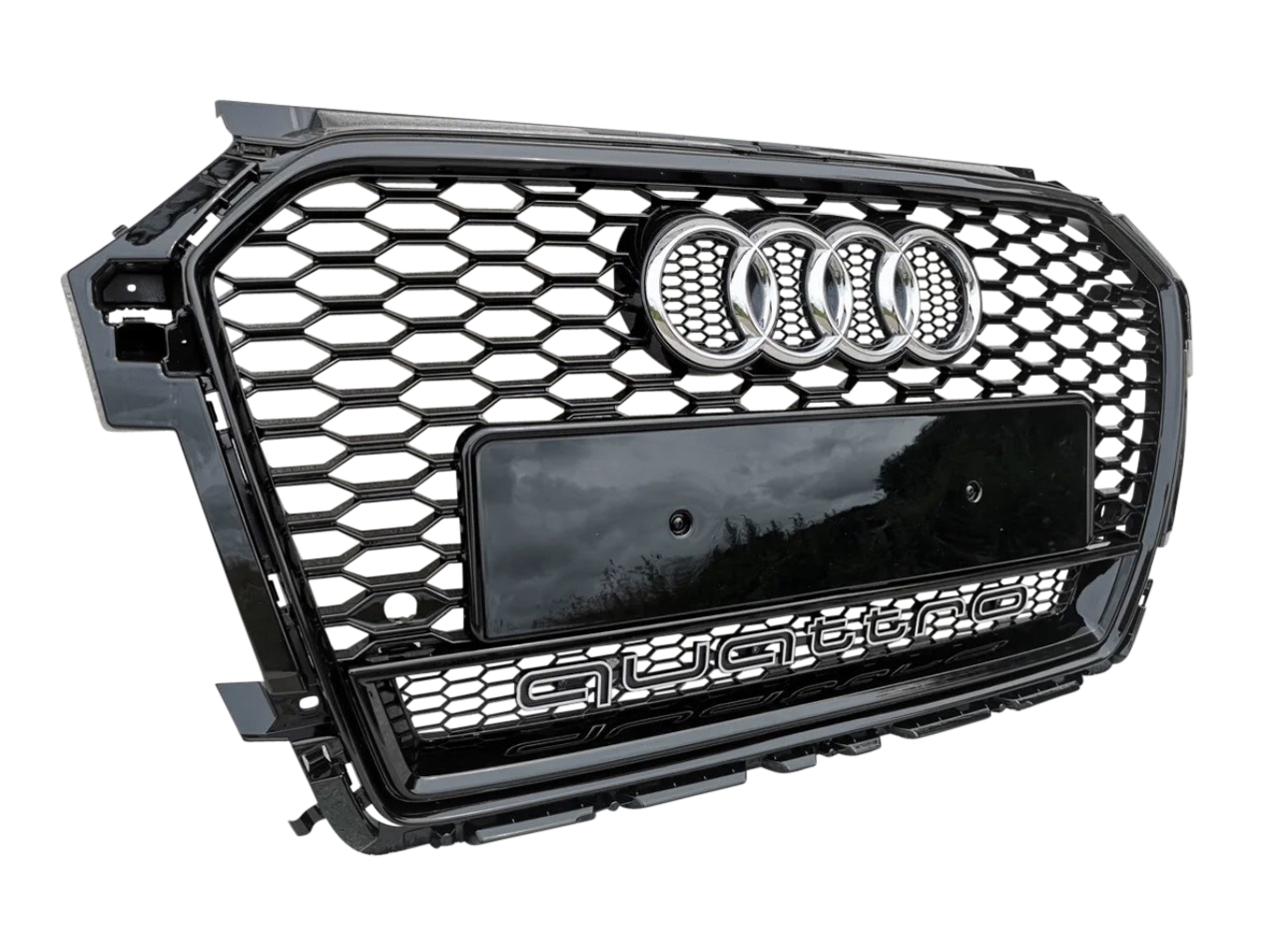 A car grille displays the Audi logo and "quattro" text in a hexagonal pattern with a glossy black finish, positioned against a light gray textured background.