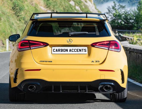 A yellow Mercedes-Benz A 35 is parked on a mountain road. It has a spoiler and visible dual exhausts. The license plate reads "CARBON ACCENTS." Lush greenery surrounds the area.