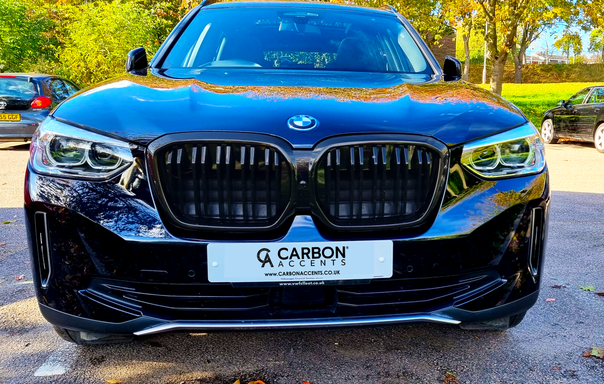 A black BMW SUV is parked on a sunlit road, surrounded by trees. The front license plate reads: "CARBON ACCENTS" with websites "CARBONACCENTS.CO.UK" and "www.rvsfleet.co.uk."