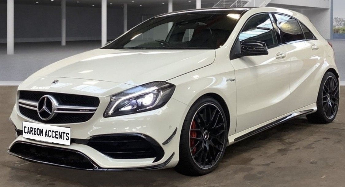 A white car with illuminated headlights is parked in an indoor showroom. It has sporty black rims and visible branding on the front. The plate reads "CARBON ACCENTS."