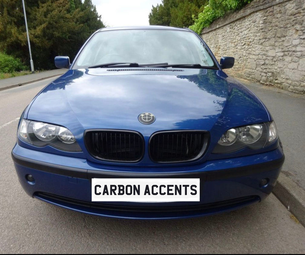 A blue sedan is parked on a street next to a stone wall. The license plate reads "CARBON ACCENTS." The vehicle's emblem is modified, surrounded by greenery and pavement.