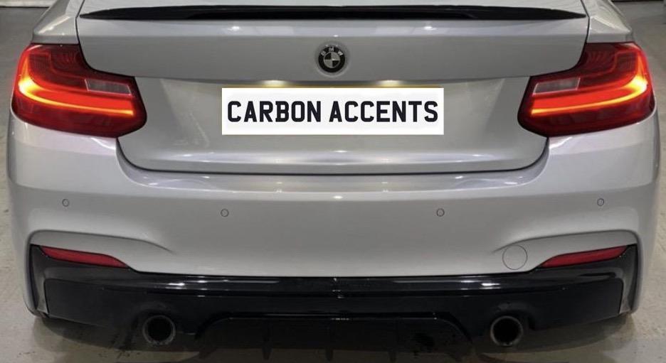 A white BMW car's rear, illuminated taillights, in a garage setting. License plate reads "CARBON ACCENTS." The car has a black lower bumper and dual exhaust pipes.