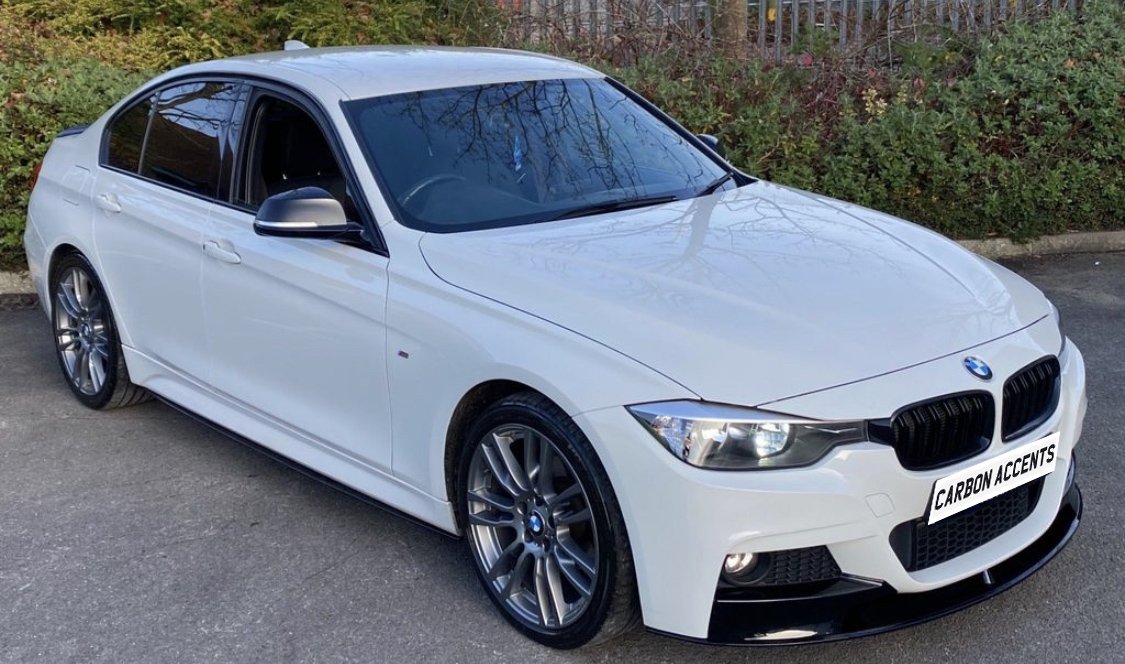 White sedan parked on asphalt, featuring sleek carbon accents and a front license plate reading "CARBON ACCENTS"; surrounded by greenery and a metal fence.