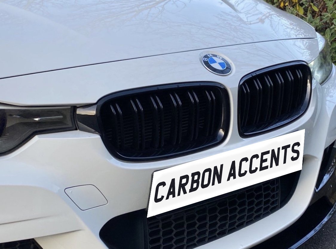 A white BMW car features a front view with black grille accents. A license plate reads "CARBON ACCENTS," set against a background of foliage.
