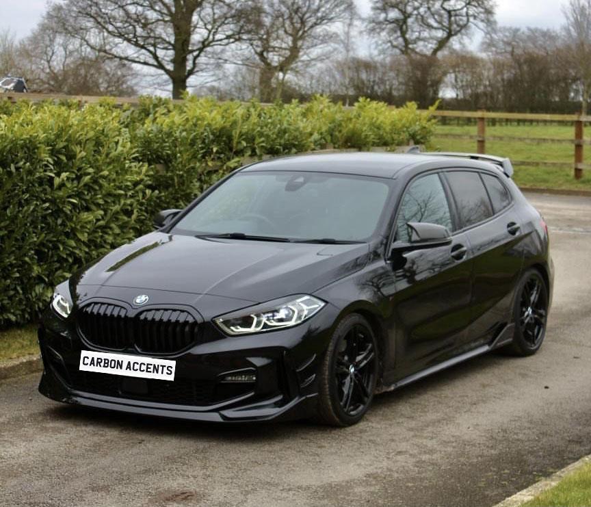 A black car is parked on a road surrounded by greenery and trees. The front license plate reads "CARBON ACCENTS."