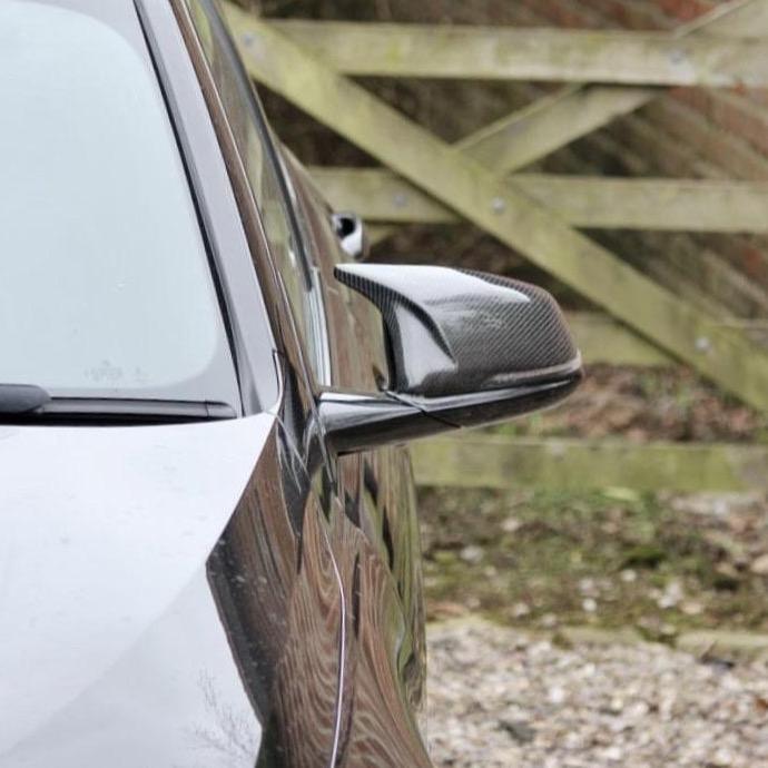 A black car's side mirror reflects a wooden gate in a gravel and grass driveway. The mirror has a carbon fiber texture and contrasts the blurred rustic background.