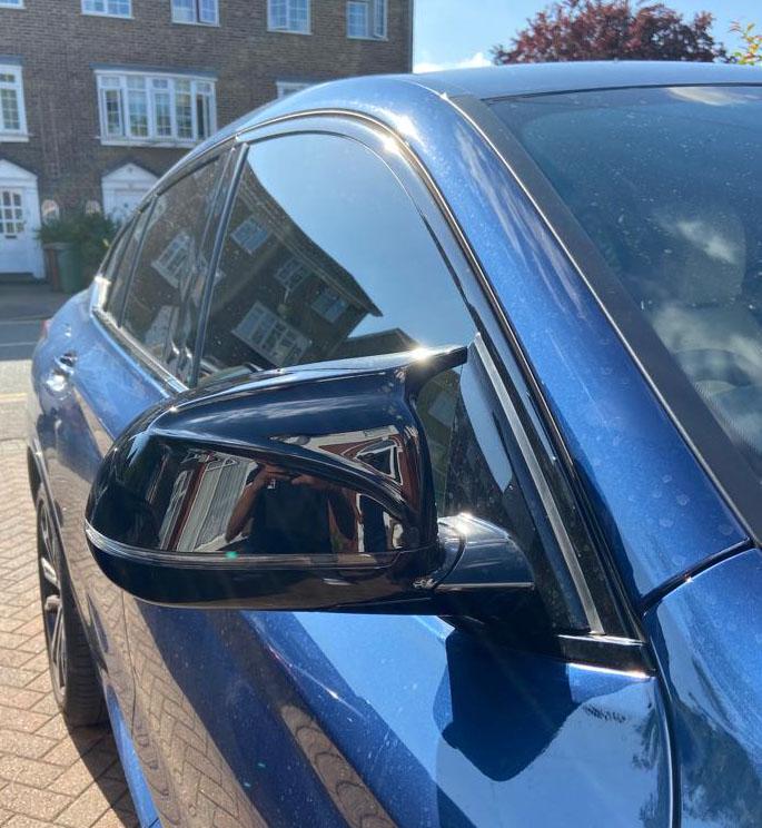 Car mirror reflects surrounding houses; blue vehicle parked on brick driveway under clear sky.