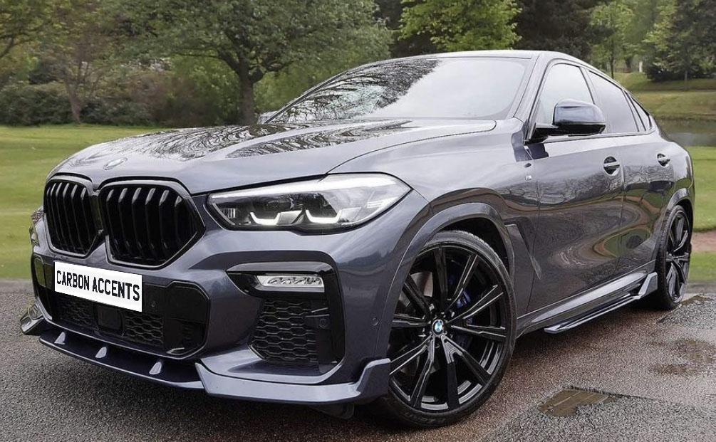 A sleek, grey SUV with large black wheels is parked on a wet road in a park-like area. The license plate reads "CARBON ACCENTS," and trees are visible in the background.