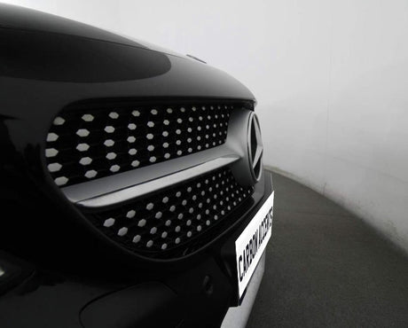 A car's front grille features a prominent logo with a pattern of hexagonal elements. It is parked in a well-lit garage. The visible text reads "CARBON ACCENTS."