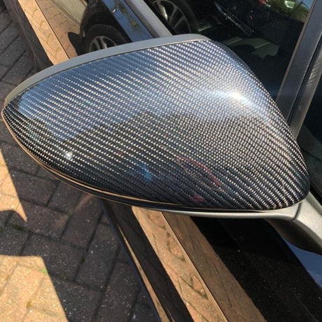 A car side mirror with a carbon fiber cover reflects light, attached to a black vehicle parked on a paved driveway, with sunlight creating a gloss effect.