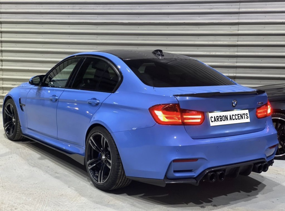 A blue BMW sedan is parked indoors, showcasing its sporty rear with illuminated taillights and carbon accents. The surroundings include a metal-paneled wall. The license plate reads "CARBON ACCENTS."