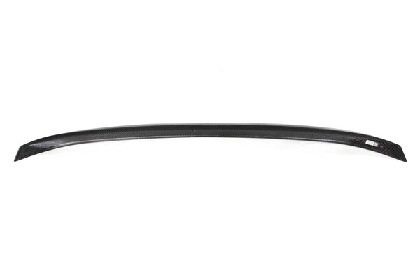 A curved, black carbon fiber car spoiler rests on a white background, displaying a smooth, textured pattern and a small label on the right side.