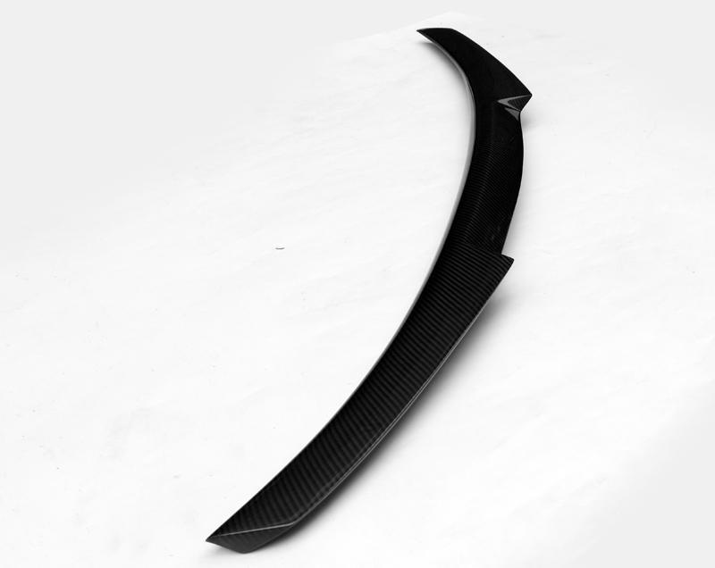 A carbon fiber car spoiler, slightly curved, rests on a plain white surface, showcasing its sleek, aerodynamic design.
