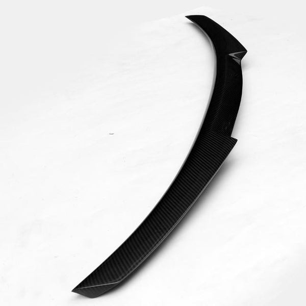 A carbon fiber car spoiler, slightly curved, rests on a plain white surface, showcasing its sleek, aerodynamic design.