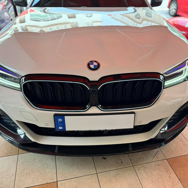 A white BMW car front is parked on a tiled floor, with reflections of buildings on the hood. Nearby are partially visible red and purple cars.