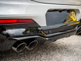 A car's rear, featuring dual exhaust pipes, is parked on asphalt. It has a glossy black diffuser and a blurred badge with a star shape on the left side.