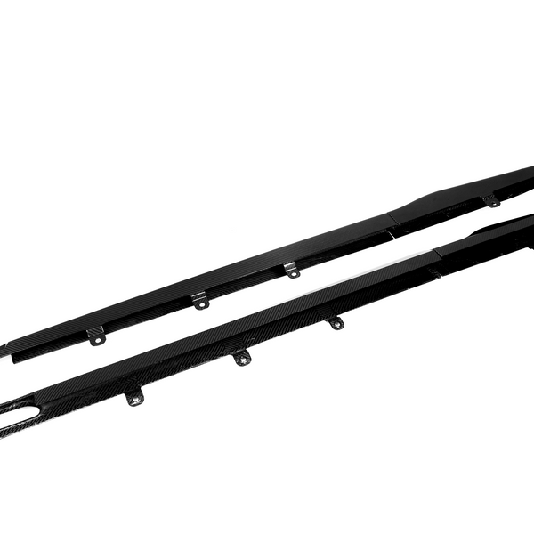 Two black carbon fiber car side skirts with attachment brackets lie against a plain white background, emphasizing their sleek design and aerodynamic purpose.