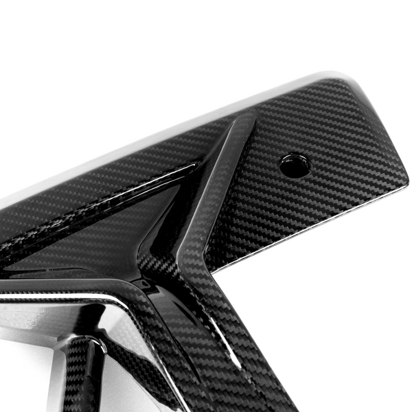 A glossy carbon fiber automotive part featuring a geometric design with sharp angles, showcased against a plain white background. The surface has a distinctive woven pattern and a small circular hole.