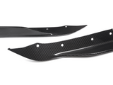 Two carbon fiber car parts, possibly splitters or fins, lie on a white background. They have aerodynamic shapes with several bolt holes for mounting.