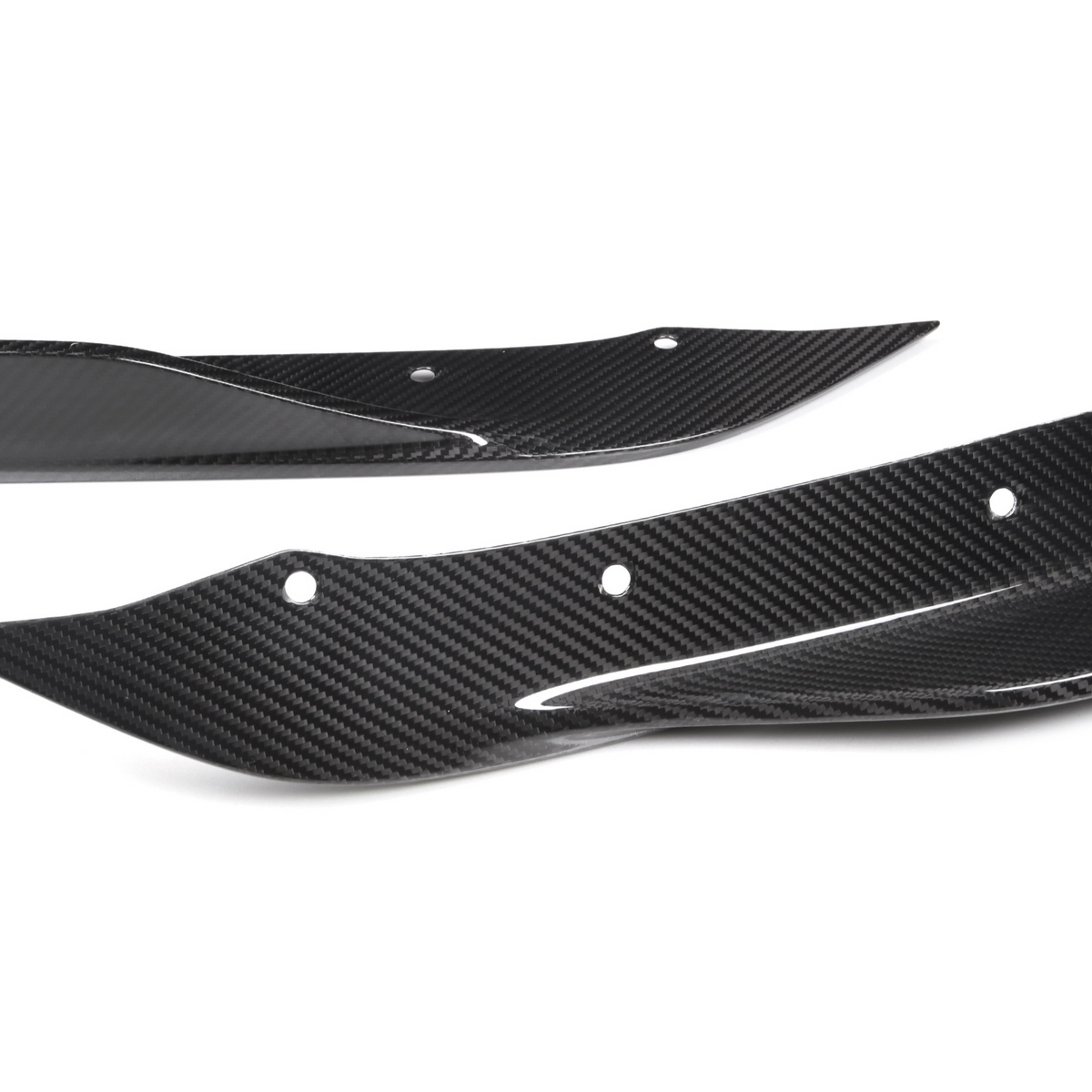 Two carbon fiber car parts, possibly splitters or fins, lie on a white background. They have aerodynamic shapes with several bolt holes for mounting.