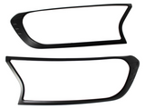 Two black plastic frames, resembling headlight covers, are positioned horizontally against a plain white background. Each frame has an asymmetric, modern design with smooth curves and sharp angles.