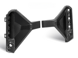 Two black carbon fiber car parts with angular designs are positioned facing each other. The left piece has visible attachment points; the right features an array of cutouts.