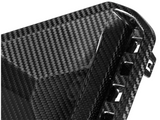 A close-up of a glossy carbon fiber object, likely a car part, with a woven pattern and angular design, featuring small vents or openings along the edge.