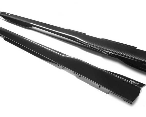 Two sleek, black carbon fiber side skirts lie parallel on a white surface. "500600" is printed on one of the skirts, adding a branded detail to enhance a car’s aerodynamic profile.