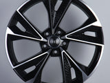 A black and silver alloy wheel with a geometric design and five dual spokes is displayed upright on a neutral background. The center features a logo with four interlocking rings.
