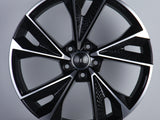 Alloy wheel with a sleek, geometric black and silver design, featuring the Audi logo in the center. Displayed against a pale background, emphasizing its modern aesthetic.