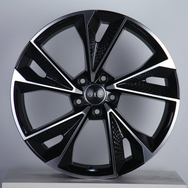 Alloy wheel with a sleek, geometric black and silver design, featuring the Audi logo in the center. Displayed against a pale background, emphasizing its modern aesthetic.