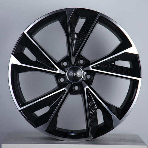Alloy wheel with a sleek, geometric black and silver design, featuring the Audi logo in the center. Displayed against a pale background, emphasizing its modern aesthetic.