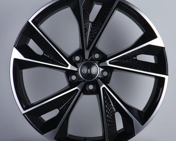 A glossy black alloy wheel, featuring a stylish multi-spoke design, stands upright against a light gray background. The center displays the Audi logo with four interlocking rings.