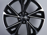 A glossy black car wheel with five split spokes features a central circle displaying four interlinked rings. It rests against a plain gray background, highlighting its modern design.