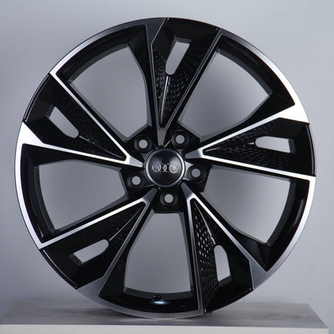 A black and silver alloy wheel, featuring a prominent logo with four interlinked circles at the center, is displayed against a plain gray background.