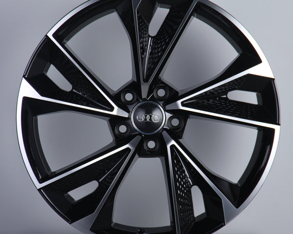 A black and silver alloy wheel is displayed upright, featuring a modern design with multiple spokes. The "Audi" logo is prominently centered. The background is a simple, neutral gray.