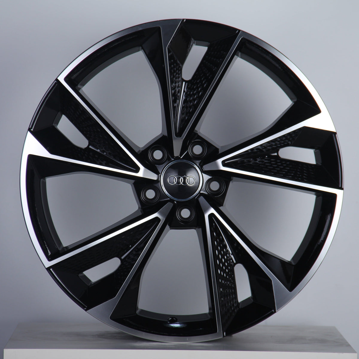 A black and silver alloy wheel is displayed upright, featuring a modern design with multiple spokes. The "Audi" logo is prominently centered. The background is a simple, neutral gray.