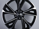 A black and silver alloy wheel with a modern, angular design is standing upright. The center features the Audi logo with interlocking rings. The background is a plain, light gray surface.