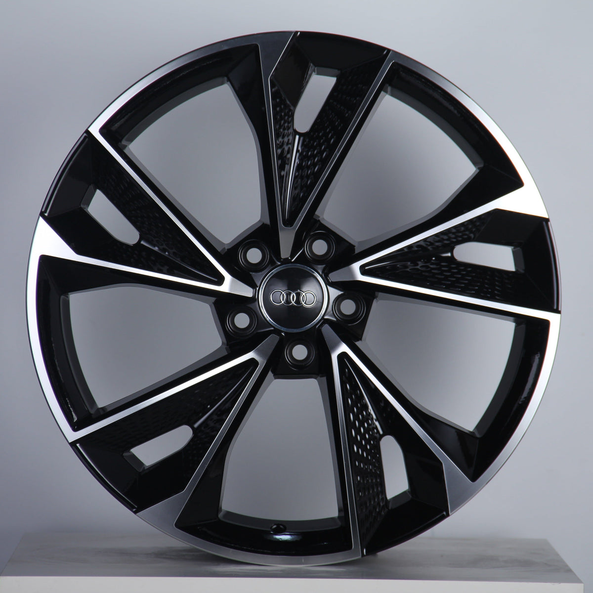 A black and silver alloy wheel with a modern, angular design is standing upright. The center features the Audi logo with interlocking rings. The background is a plain, light gray surface.
