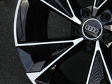 A stylish, glossy black-and-silver car wheel with circular Audi logo, featuring geometric design and polished surface, rests on an asphalt surface.