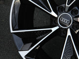 A polished black and white car wheel rim features the Audi logo at the center. It is resting on a rough, gray asphalt surface.