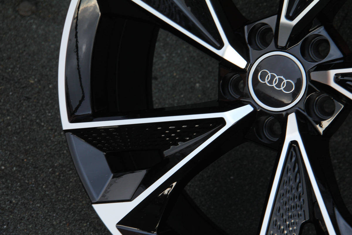 A close-up of an Audi car wheel displays a glossy black and silver design with the Audi logo in the center, resting on a textured asphalt surface.