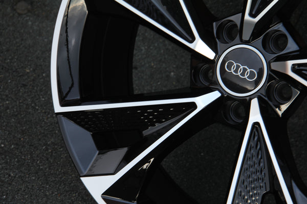 A shiny black and silver car wheel rim with the Audi logo at the center, positioned on a dark, textured ground surface.