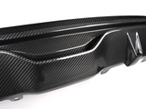 A carbon fiber car bumper diffuser features a textured design, showcasing glossy, aerodynamic curves and fins, mounted on a white surface, emphasizing its sleek automotive styling.