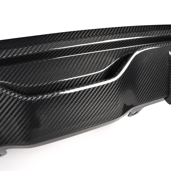 A carbon fiber car bumper diffuser features a textured design, showcasing glossy, aerodynamic curves and fins, mounted on a white surface, emphasizing its sleek automotive styling.