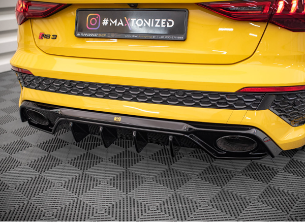 A yellow car's rear bumper, featuring dual exhausts and a honeycomb pattern, sits on a checkered floor. Text on a sign reads, “#MAXTONIZED” and contact details for "TRZONEKON SPOJ.”