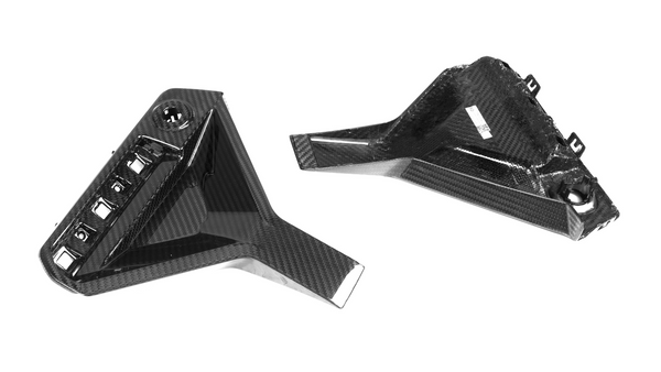 Carbon fiber components lying on a white surface; one has a rectangular grid pattern, and the other features mounting clips and a small label.