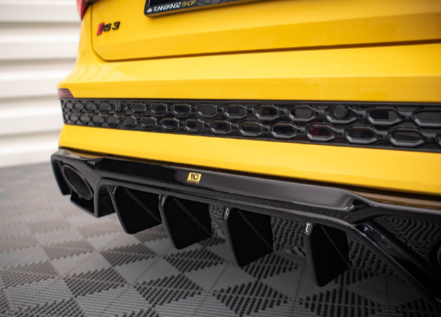 A yellow car with "RS3" badge features a sleek black rear diffuser. The car is stationary on a checkered garage floor, showcasing its sporty rear design.